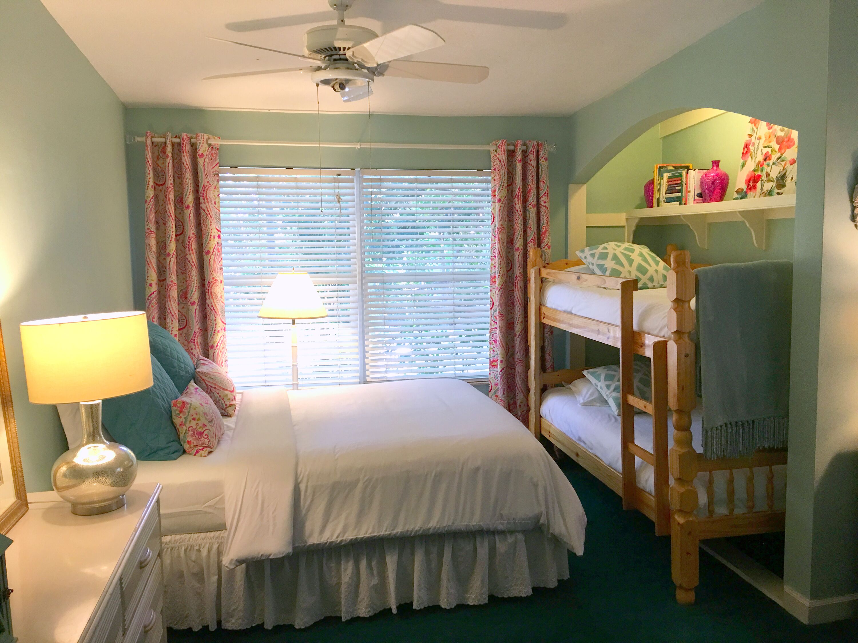 A bedroom with a bed, a nightstand, a lamp, a ceiling fan, a window with curtains, and a bunk bed.