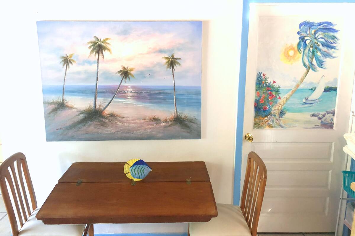A dining area with a wooden table and chairs, a painting of a beach scene, and a door with a tropical-themed painting.
