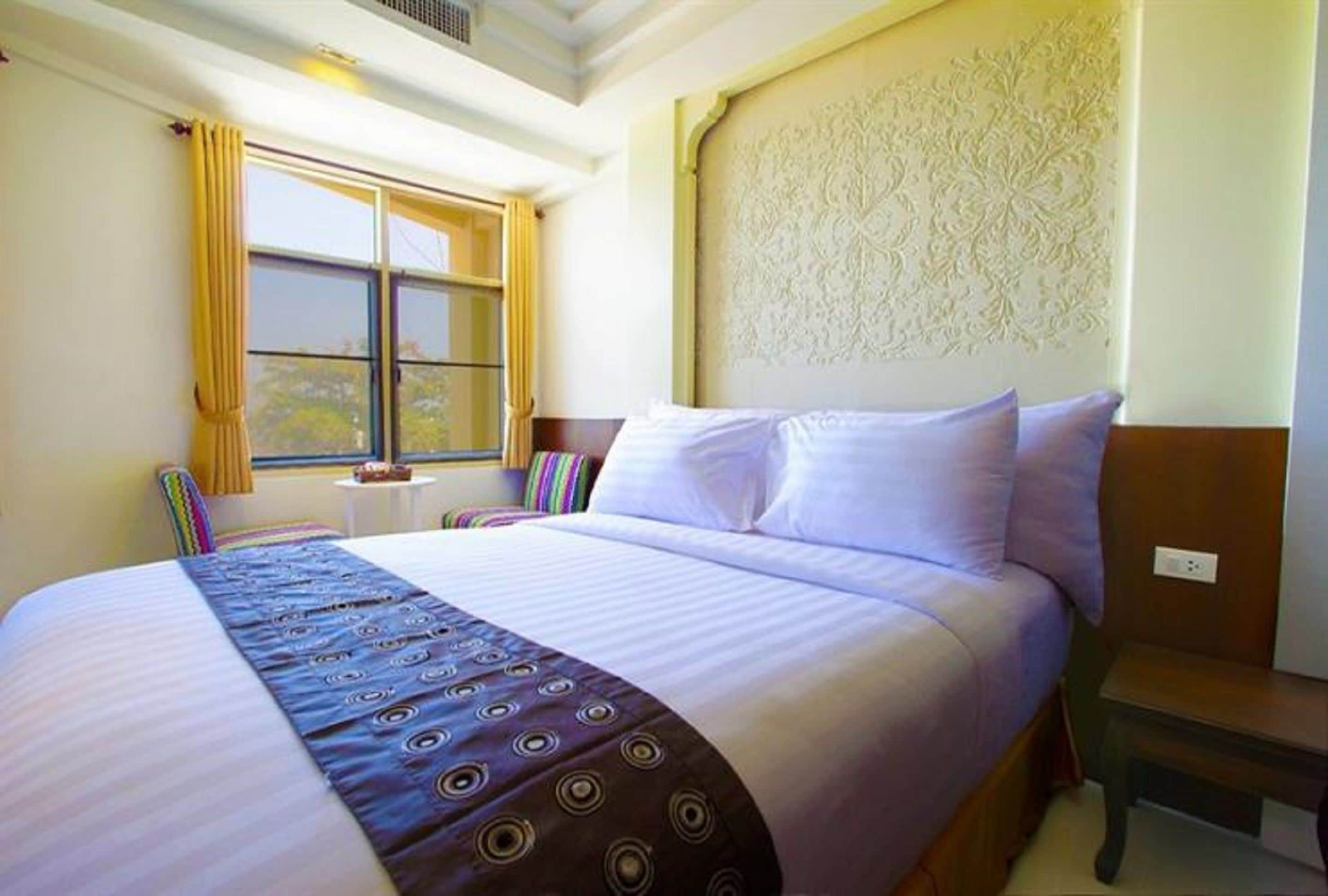 A hotel room with a large bed, a window with yellow curtains, a small table with a lamp, and a decorative headboard.