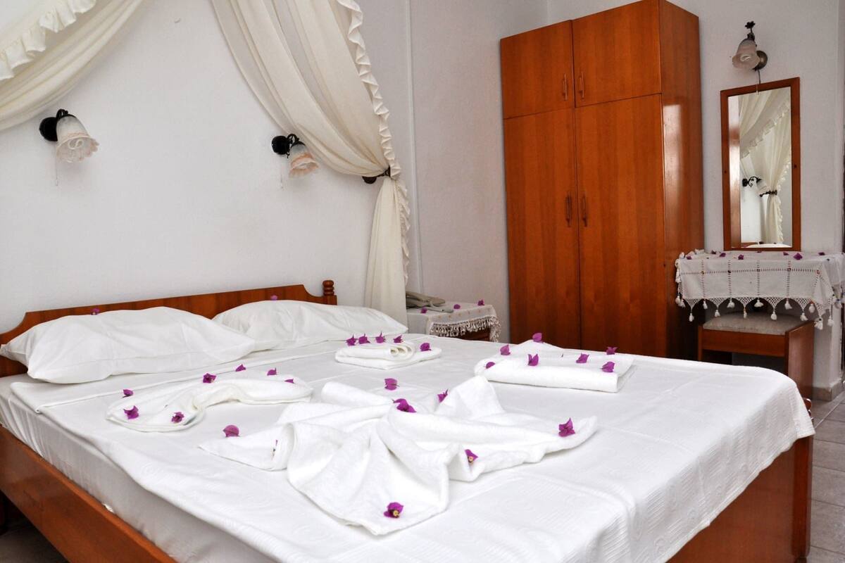 A neatly made bed with white linens and purple flower petals, a wooden wardrobe, a bedside table with a lamp, and a mirror on the wall.