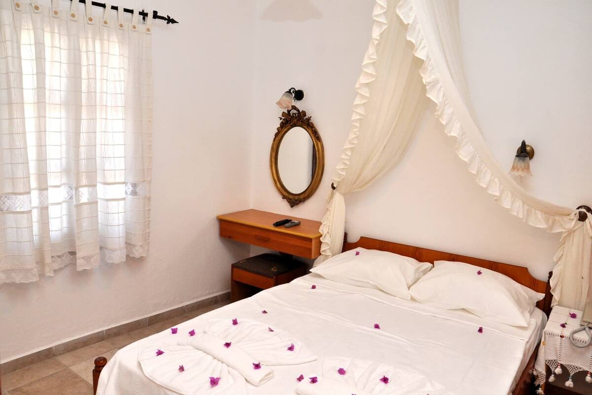 A neatly made bed with white linens and purple flower petals, a wooden nightstand with a remote control, a wall-mounted mirror, and sheer curtains.