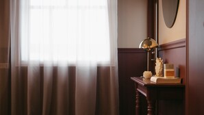 Une pièce dotée d’une grande fenêtre ornée de rideaux légers, d’un bureau en bois avec une lampe et d’un miroir accroché au mur.