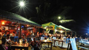 An outdoor dining area with tables and chairs, a building with a tiled roof, and palm trees in the background. - Sana Guest (Weligama)