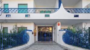 Entrada de un edificio con una verja metálica azul, un camino de baldosas y fachada blanca.