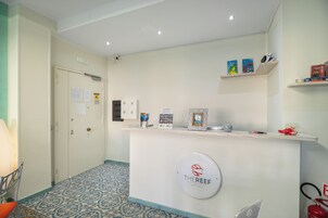 Un'area reception con un banco che mostra un logo, un estintore e un tubo antincendio. Si vedono scaffali con libri e oggetti decorativi, oltre a un pavimento a motivi.
