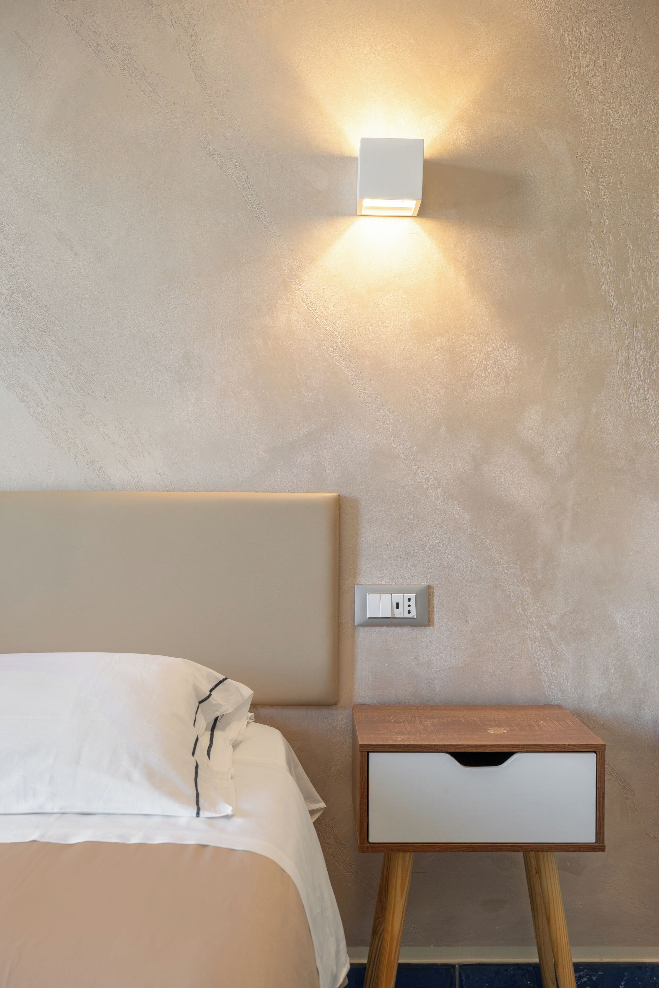 A neatly made bed with white linens, a bedside table with a drawer, and a wall-mounted light fixture.