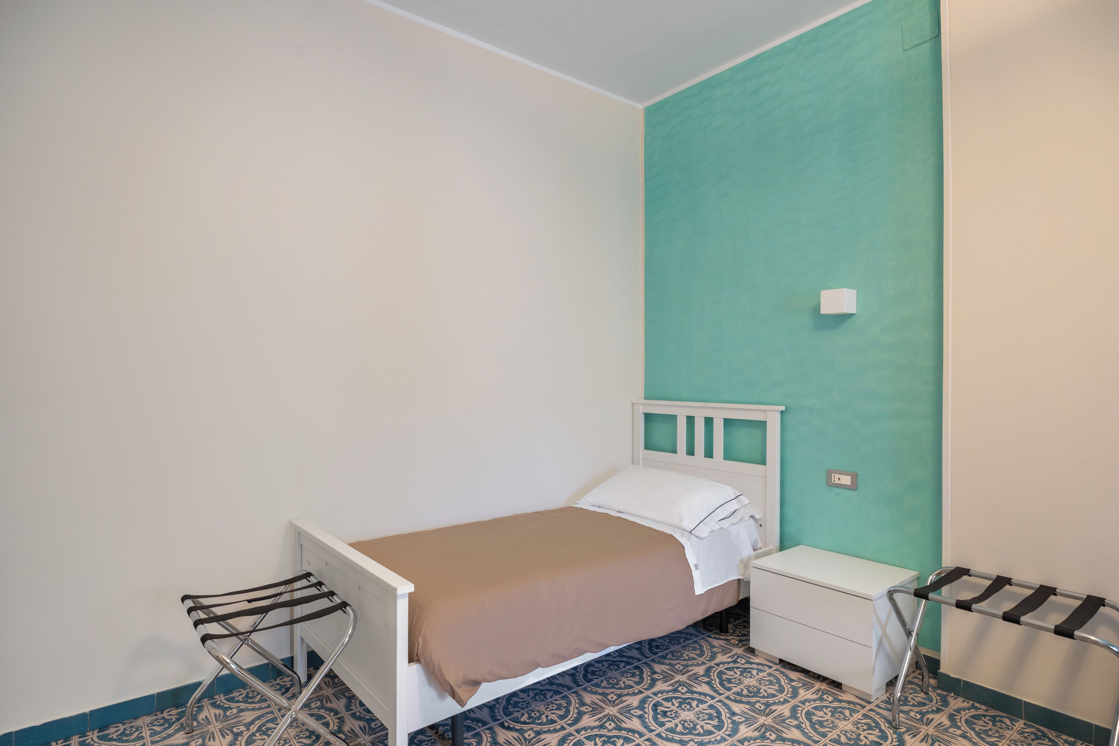 A small, single-bed room with a white headboard, a brown bedspread, and a white nightstand. The walls are white and one is teal. There is a patterned blue and white floor.