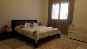 Un dormitorio con una cama, dos lámparas de noche, una cuna y una ventana con cortinas.