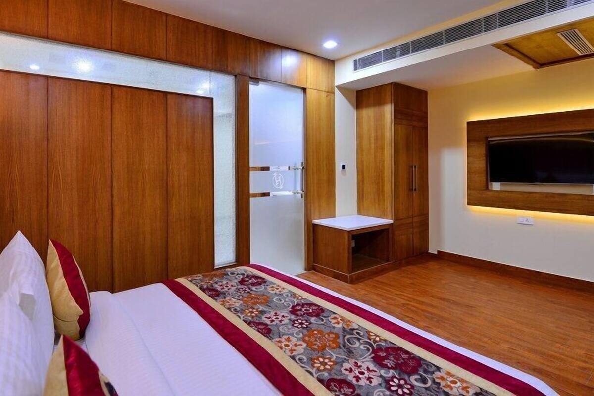 Una habitación de hotel con cabecera de madera, cama con ropa de cama blanca y roja, televisor montado en la pared y un pequeño mueble de madera.