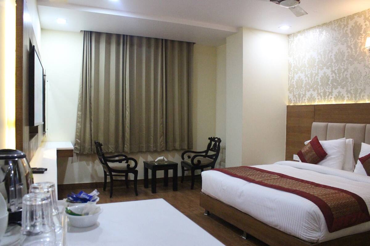 Una habitación de hotel con cama, escritorio con silla, una mesita y televisión en la pared.