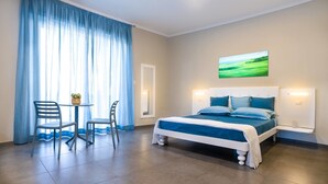 A bedroom with a bed, a desk, two chairs, a window with curtains, a wall-mounted light, and a painting. - La Bouganville (Vibonati)
