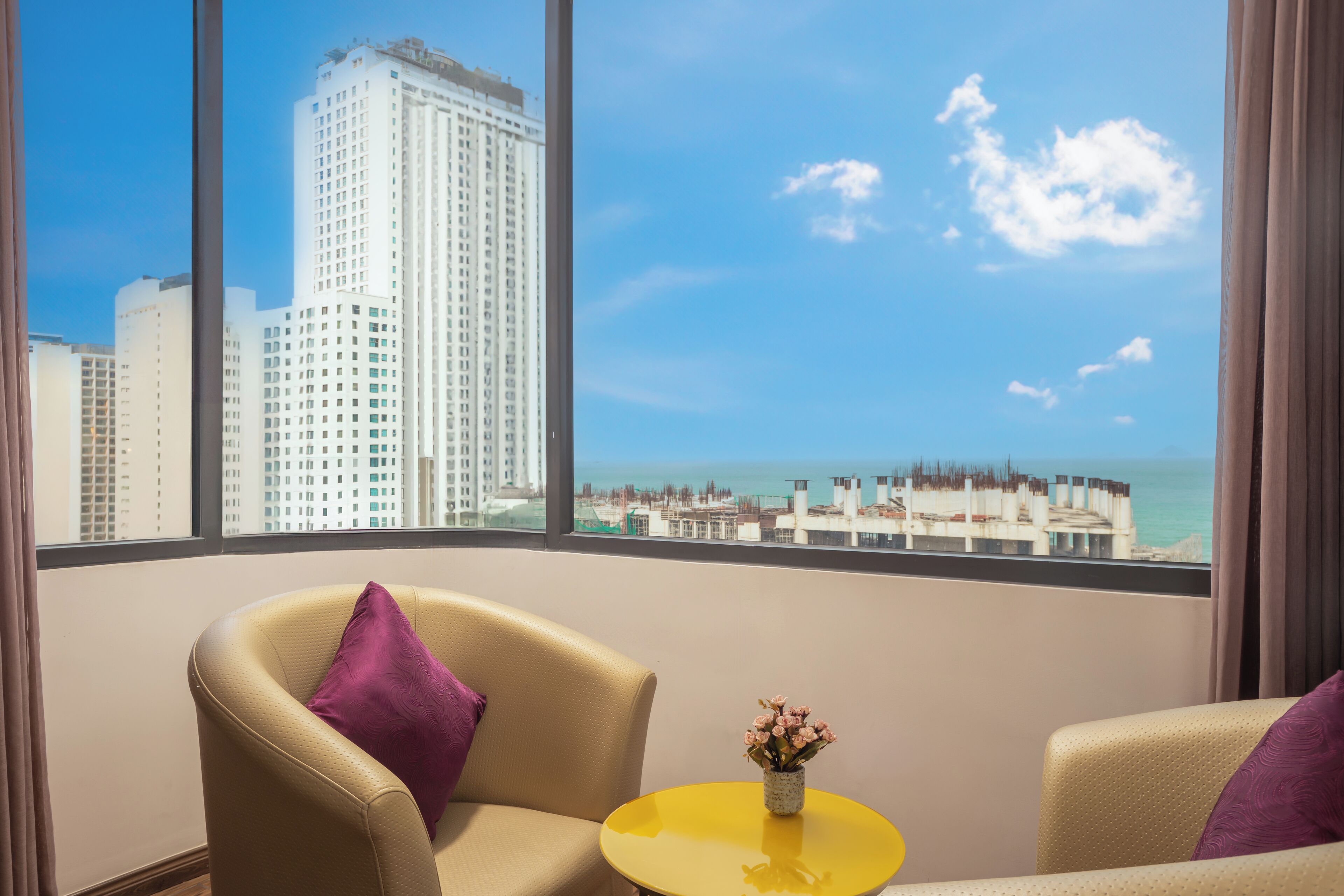 A hotel room with a large window offering a view of a tall building and a beach. There are two chairs and a small yellow table with a vase of flowers.