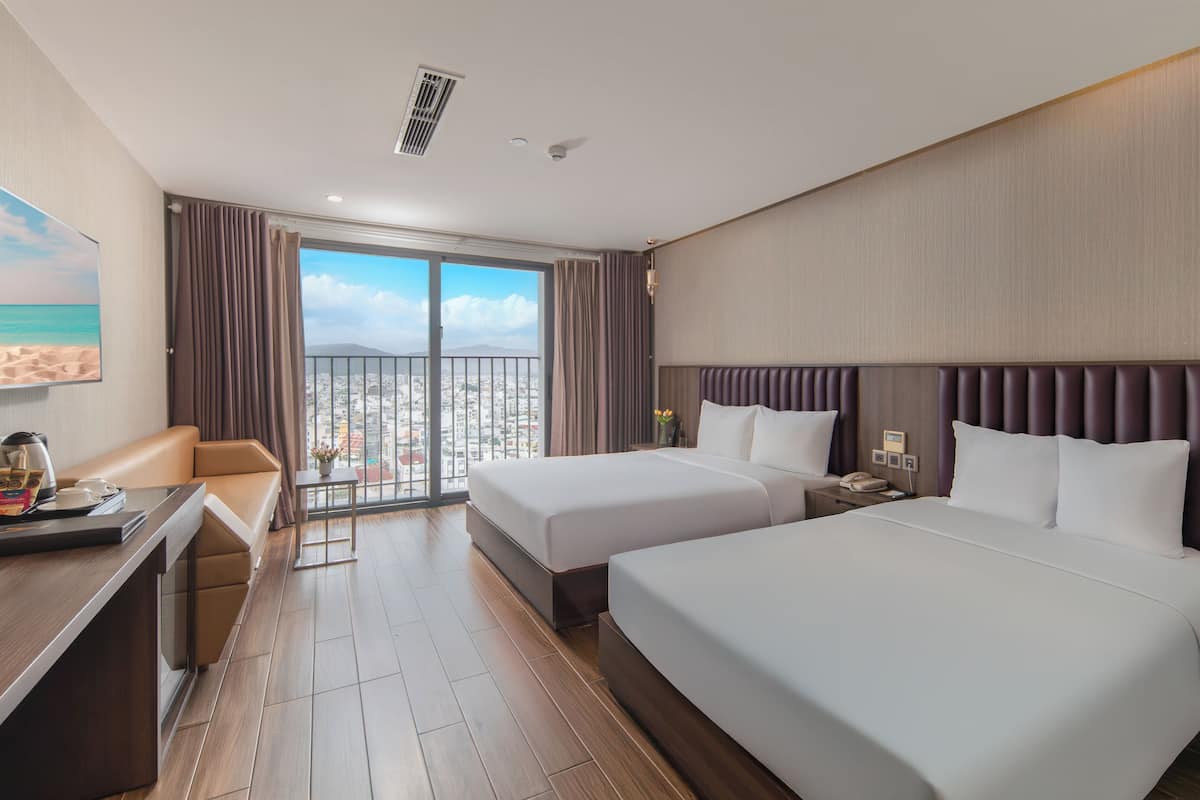 A hotel room with two beds, a desk, a chair, and a balcony with a city view.