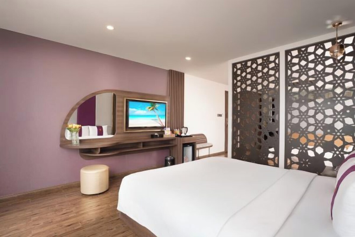 Habitació d'hotel moderna amb un llit gran, una televisió penjada en un panell de fusta, una partició decorativa i un petit tamboret.