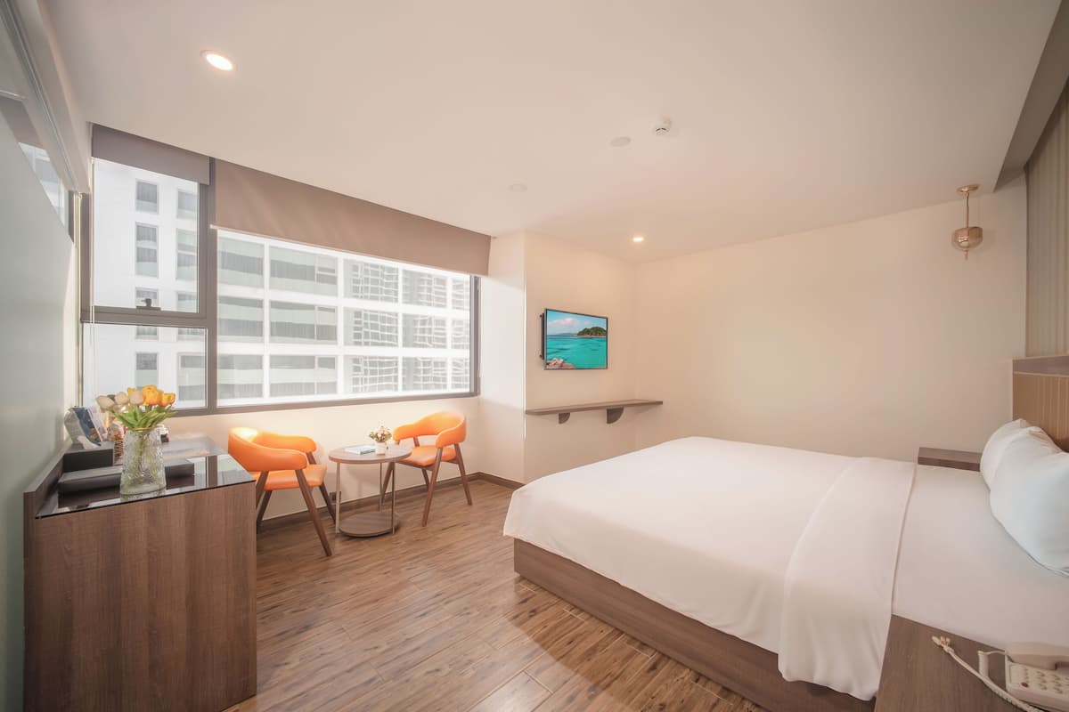 A modern hotel room with a large bed, a desk with a vase of flowers, two orange chairs, a small table, and a large window with a view of buildings.