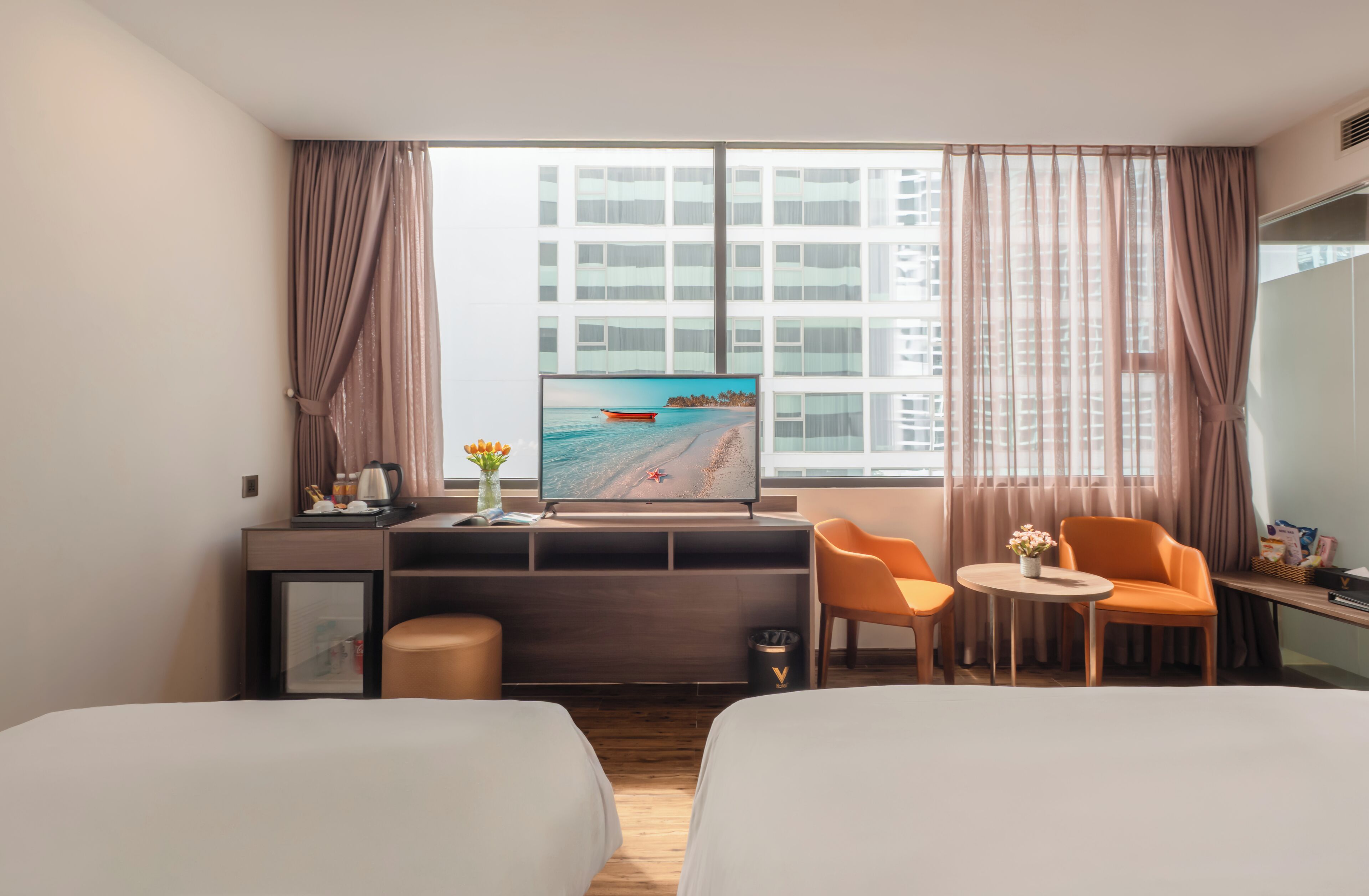 A hotel room with two beds, a desk with a TV displaying a beach scene, a small table with a vase, and a window with curtains.