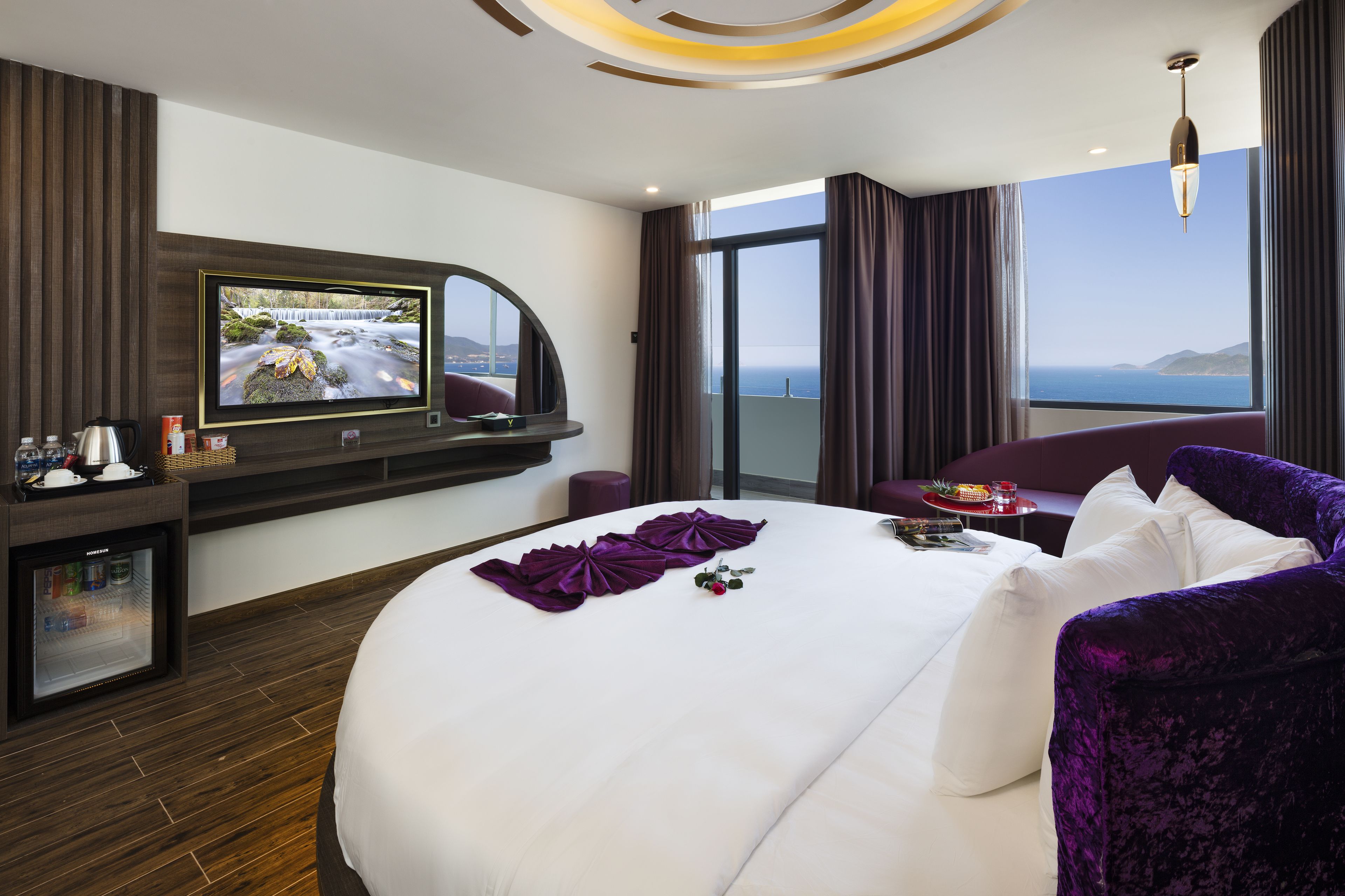 A modern hotel room with a large bed, a flat-screen TV, a balcony with a sea view, and a minibar.
