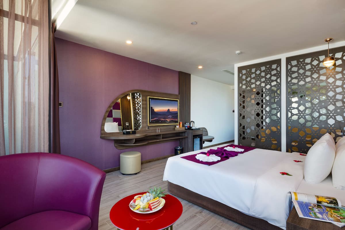 A hotel room with a bed, a red table with fruit, a purple armchair, a TV, and a decorative partition.