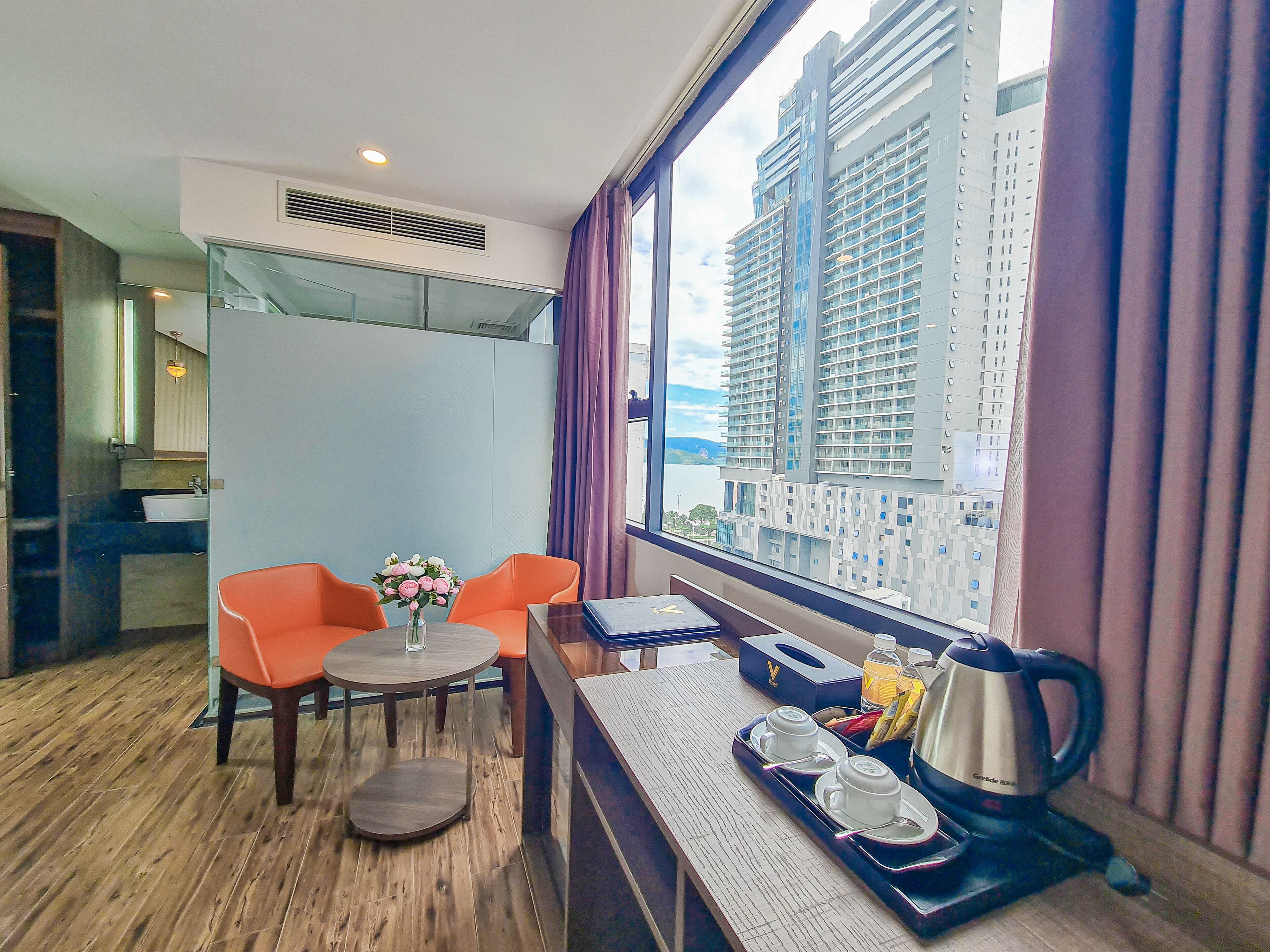 Habitación de hotel con un ventanal grande, una mesa de comedor con sillas naranjas, una mesita redonda con un jarrón de flores y una bandeja con juego de té.
