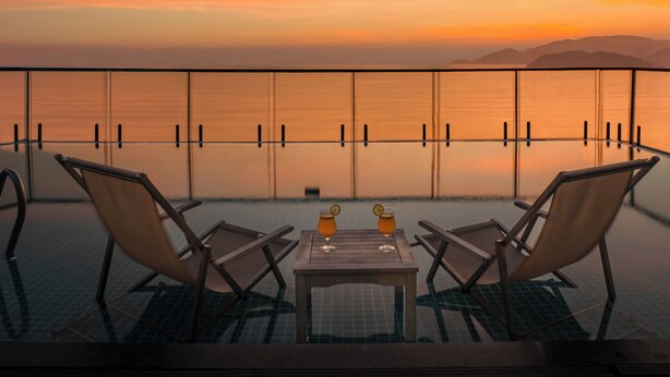 Piscina infinita com duas espreguiçadeiras e uma mesa, com vista para um pôr do sol sobre o oceano.