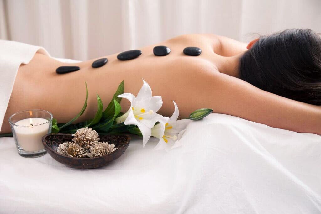 En person får massage med svarta stenar på ryggen, omgiven av vita blommor och ett ljus.