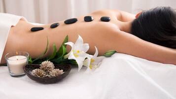 A person receiving a massage with black stones on their back, surrounded by white flowers and a candle.