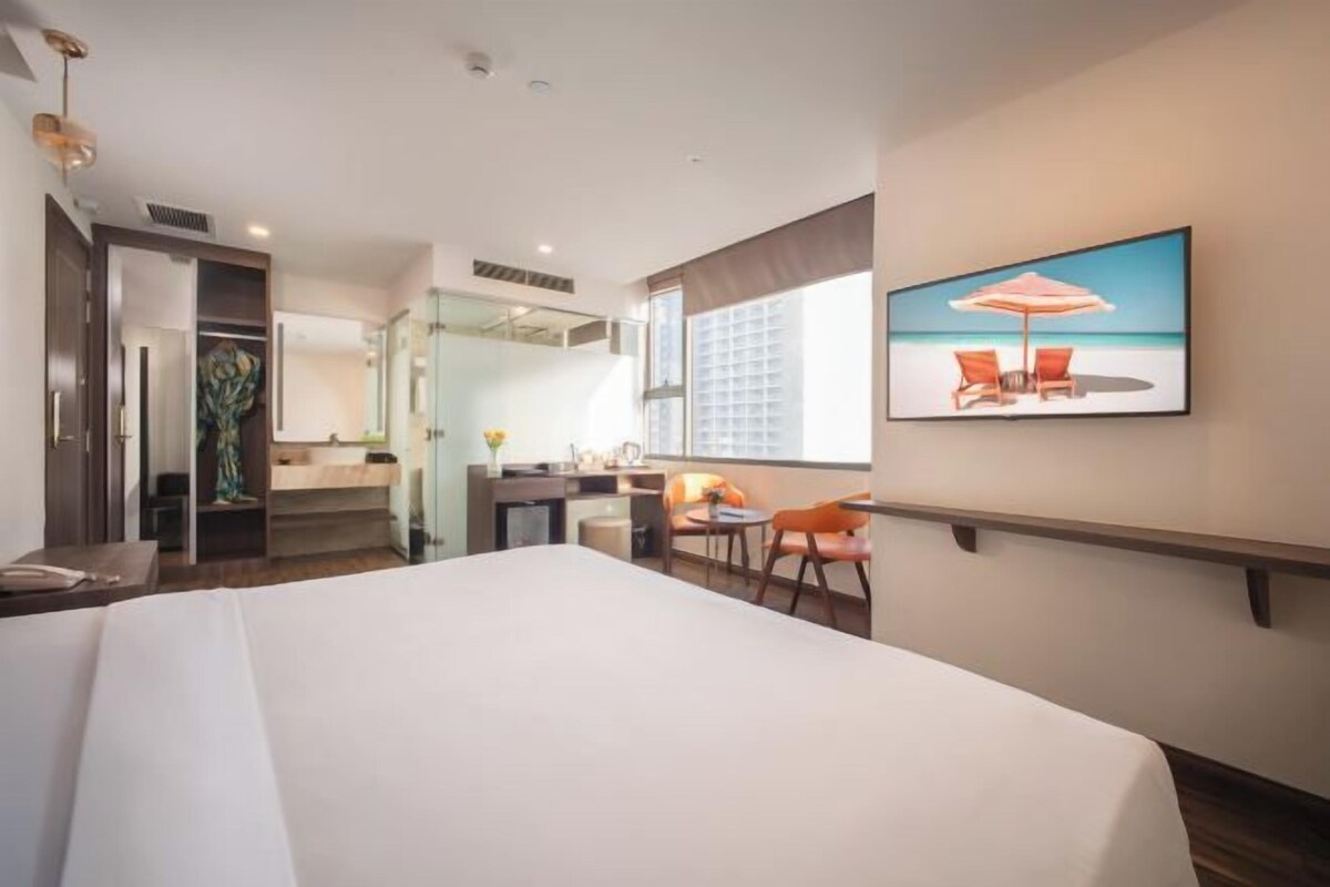 A hotel room with a large bed, a desk with a chair, a small table with two chairs, a window with a view of the beach, and a painting of a beach scene.