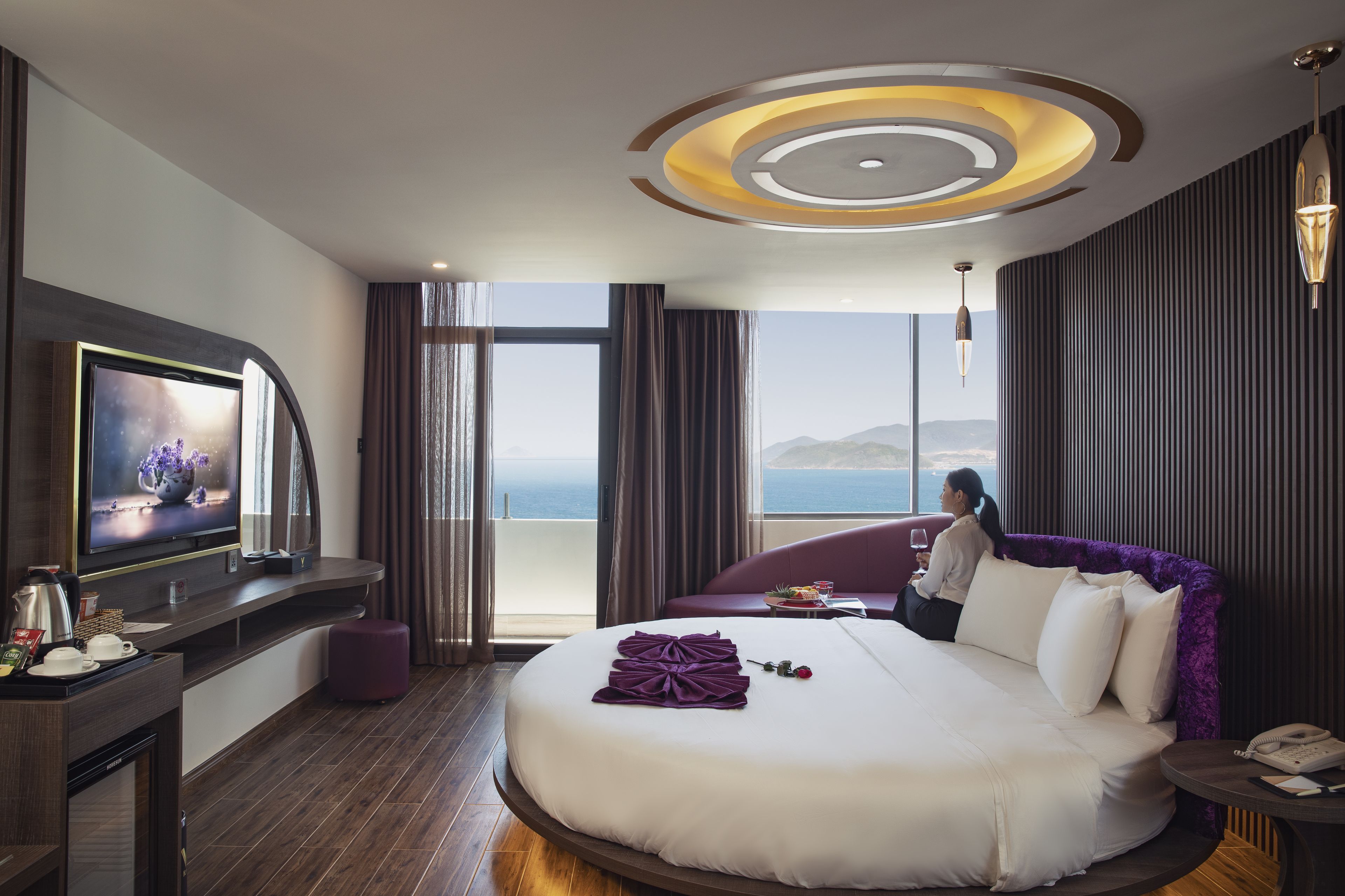 Ein modernes Hotelzimmer mit einem großen Bett, einem Fernseher auf einer gebogenen Konsole, einem Balkon mit Meerblick und einem stilvollen Deckenstil.