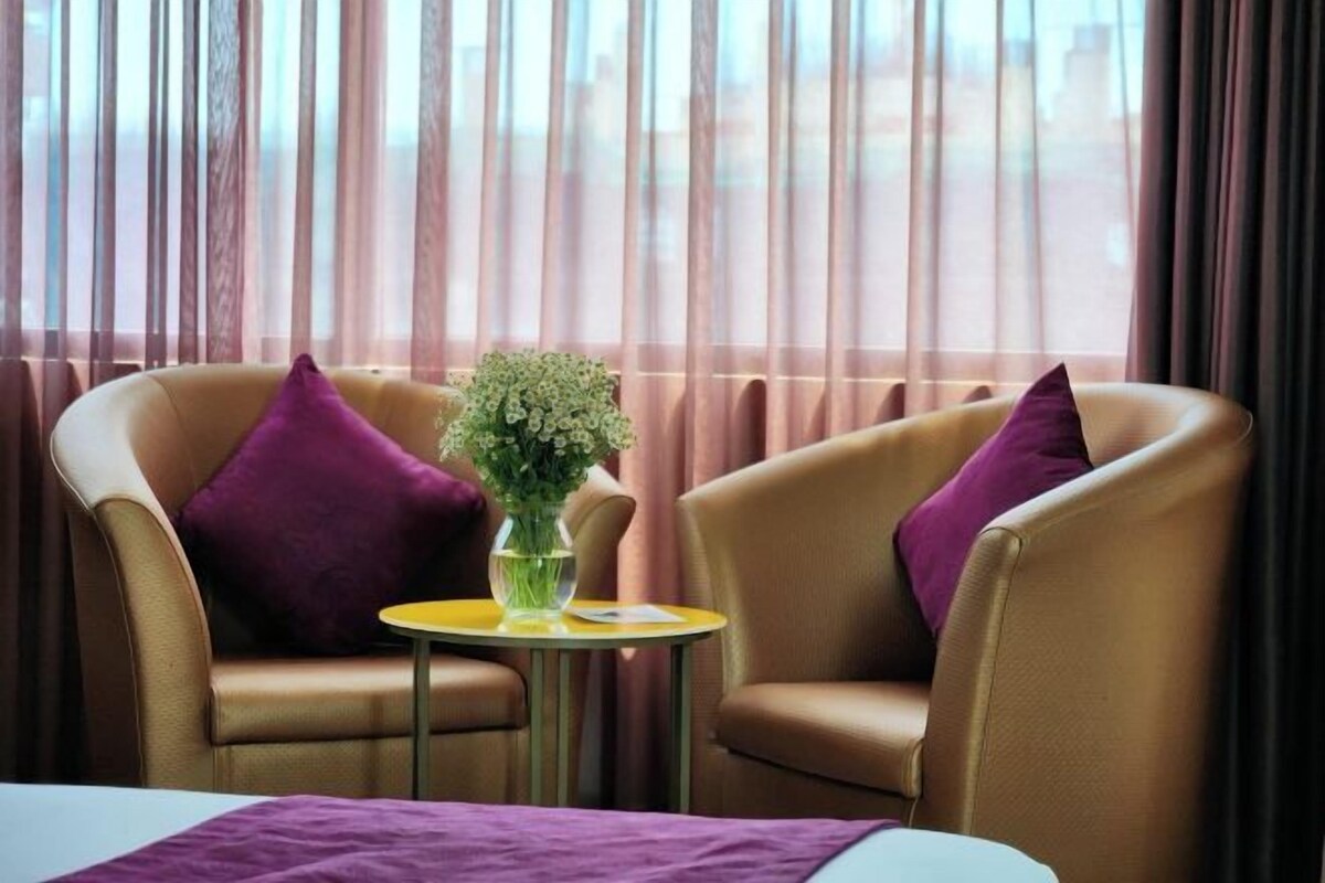 A hotel room with two leather armchairs, a small round table with a vase of flowers, and a bed with a purple blanket.