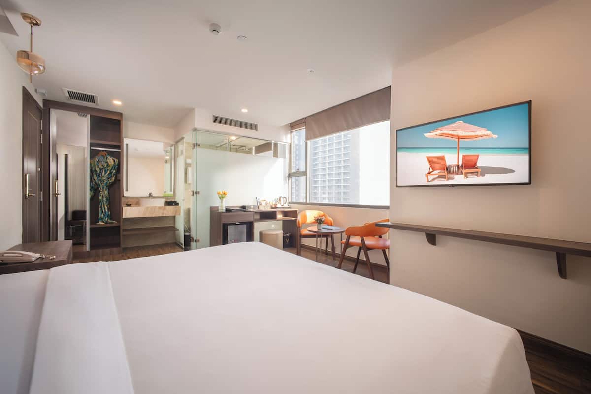 A hotel room with a large bed, a desk with a chair, a small table with a chair, a TV on the wall, and a window with a beach view.