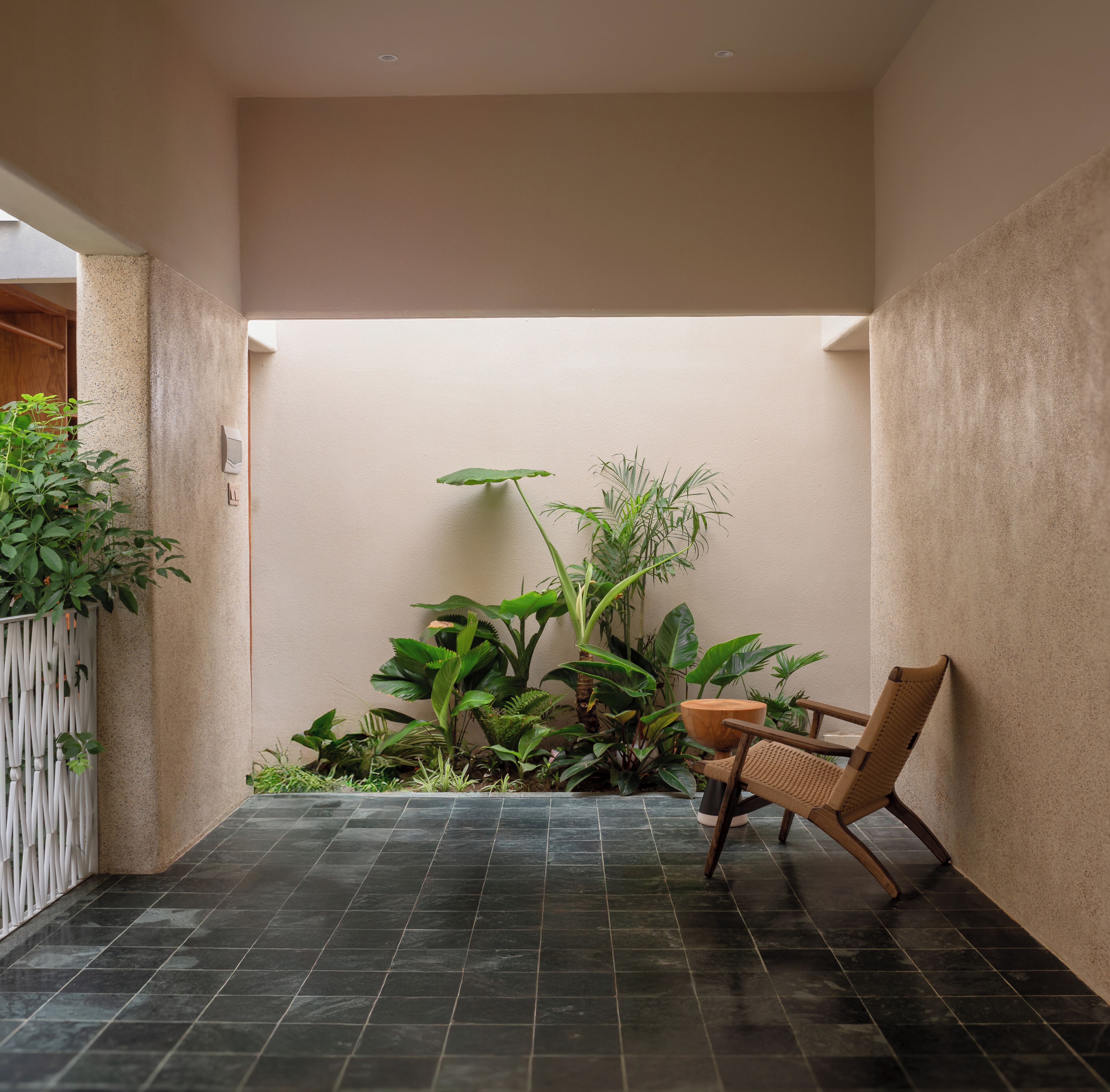Un piccolo patio recintato con pavimento piastrellato, una sedia di legno e diverse piante verdi.