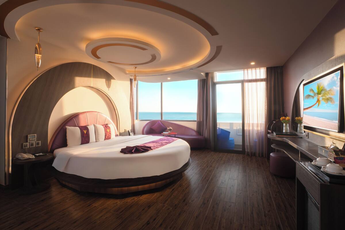 A modern hotel room with a large bed, a view of the sea, and a TV on the wall.