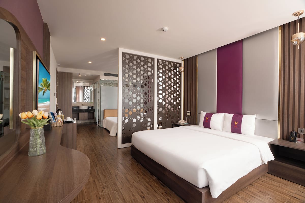 A modern hotel room with a large bed, a curved wooden desk, a vase of flowers, and a view of the bathroom and kitchen through a decorative screen.