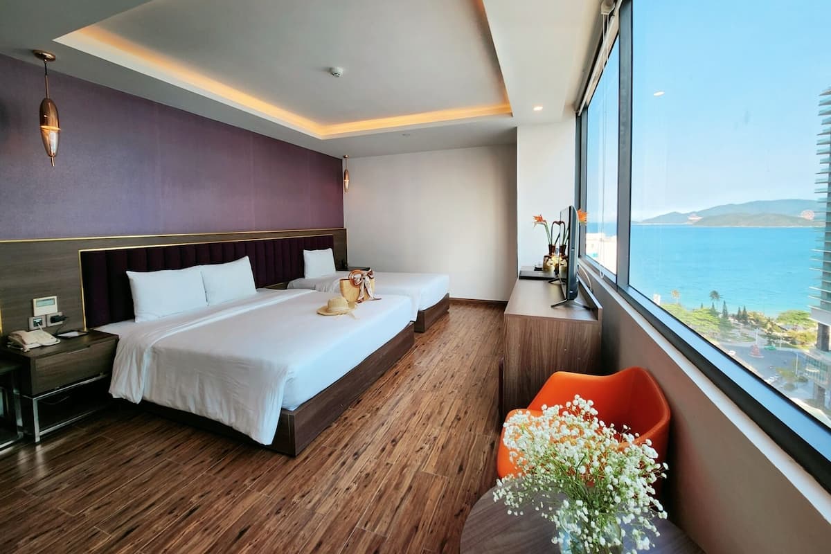 A modern hotel room with two beds, a large window with a view of the sea, and a small table with flowers.