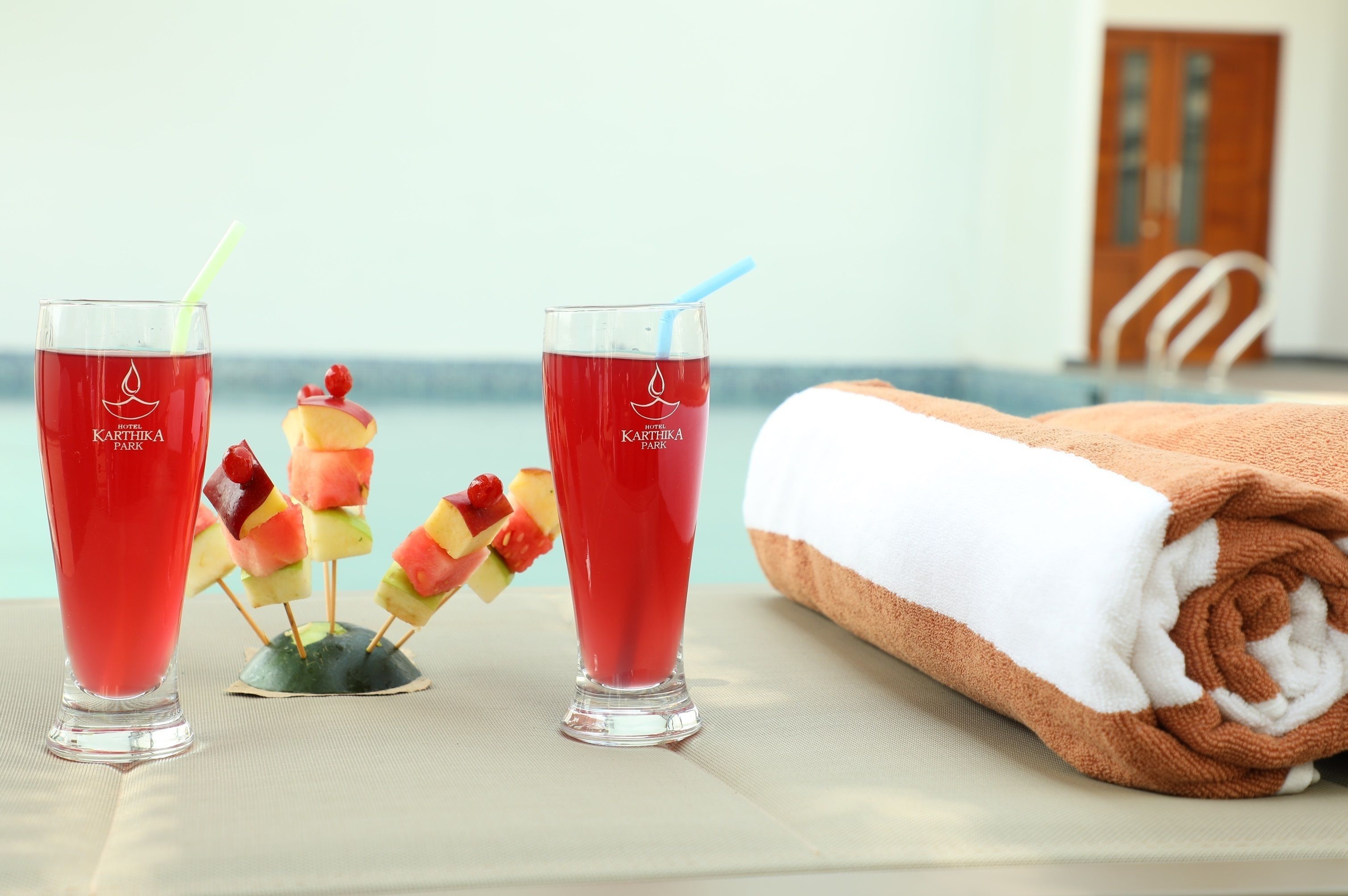 Zwei Gläser rotes Getränk mit Trinkhalmen, ein Handtuch und Obstspieße auf einem Tisch am Pool.