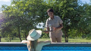 Une scène se déroulant au bord d’une piscine, où l’on voit une personne debout près de la piscine, tenant un plateau sur lequel se trouvent un verre et une bouteille.