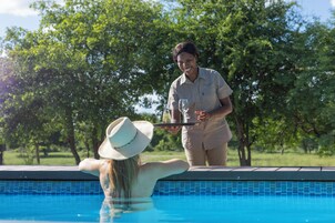 A poolside scene with a person standing by the pool, holding a tray with a glass and a bottle.