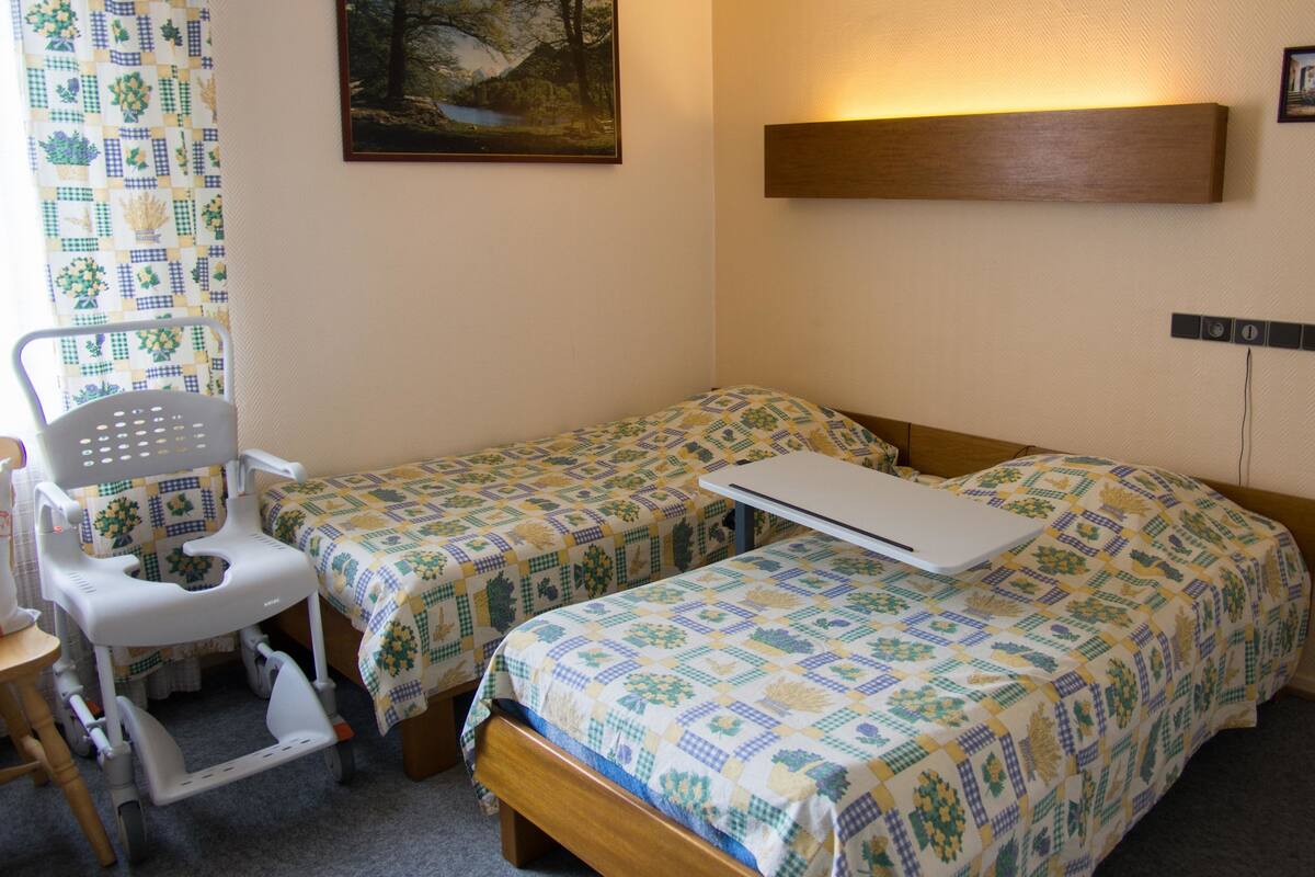 A room with two beds, a wheelchair, and a painting on the wall.