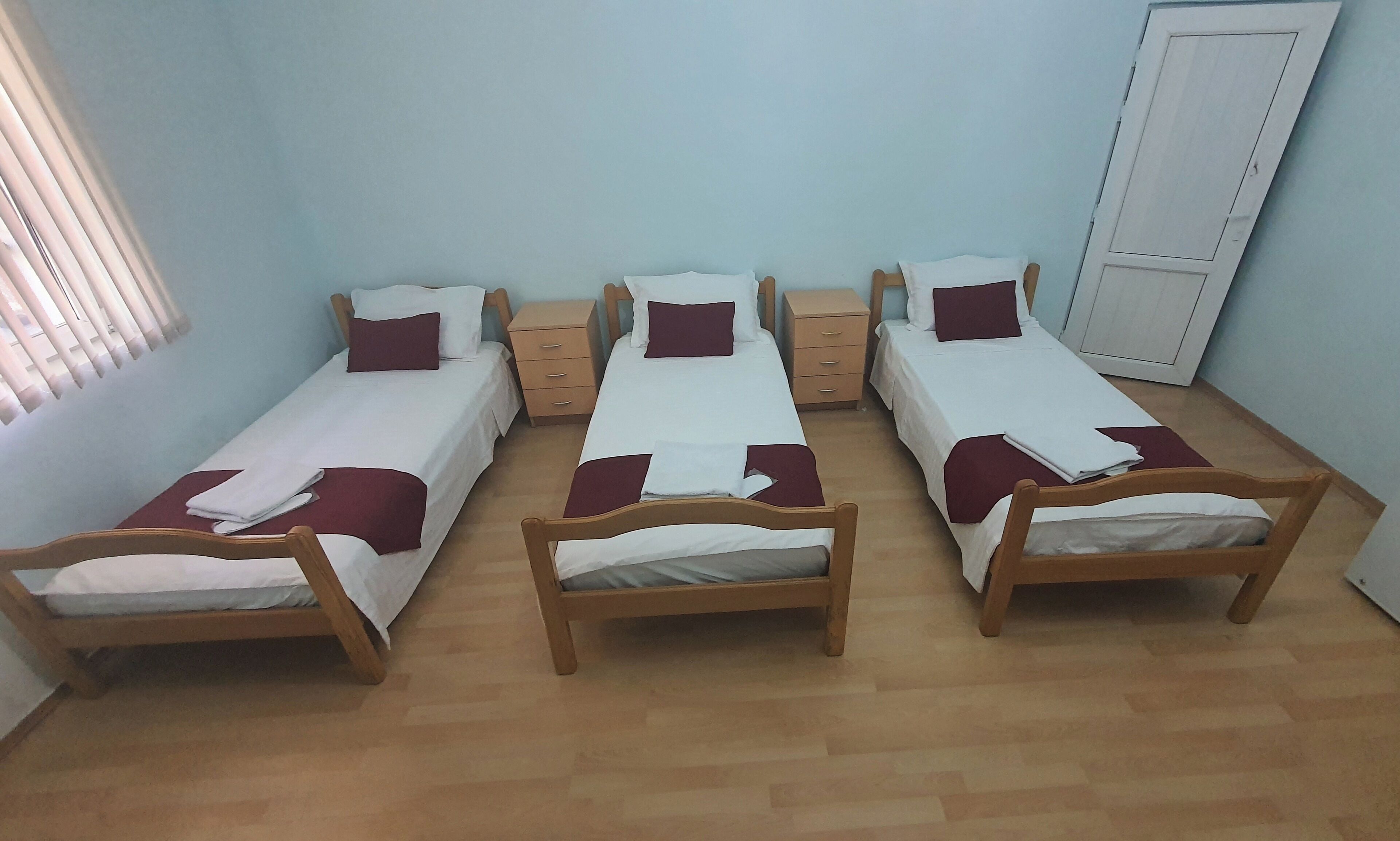 A room with three single beds, each with a wooden frame and white bedding. There are two bedside tables with drawers, and a wardrobe against the wall.