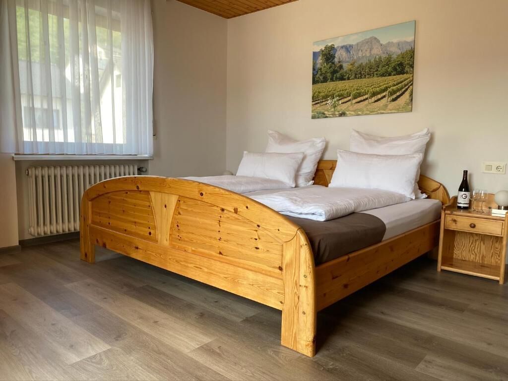 Una cama de madera con ropa blanca en una habitación con radiador, una ventana con cortinas traslúcidas y un cuadro en la pared.