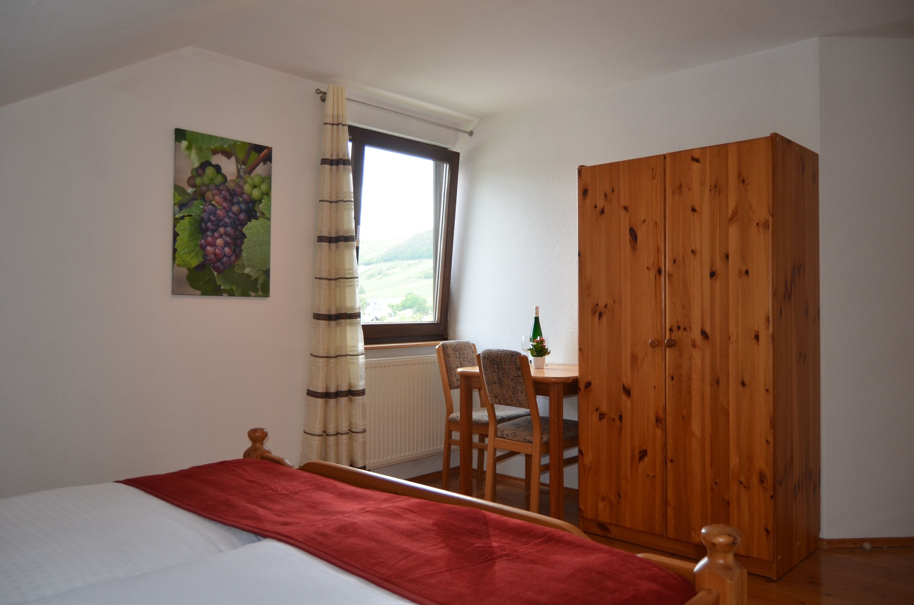 A bedroom with a bed, a wooden wardrobe, a window with a view, a table with a bottle, and a chair.
