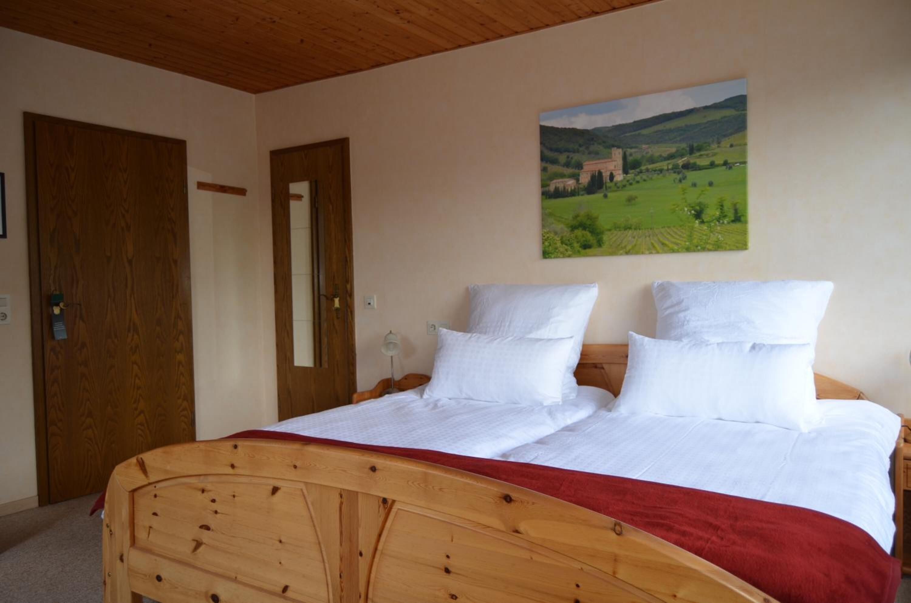 A wooden bed with white bedding and a red blanket, a wooden door, and a landscape painting on the wall.