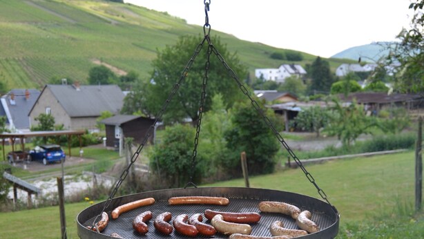 Ein Grill mit Bratwurst und ein Feuerkorb in einer grünen, offenen Umgebung.