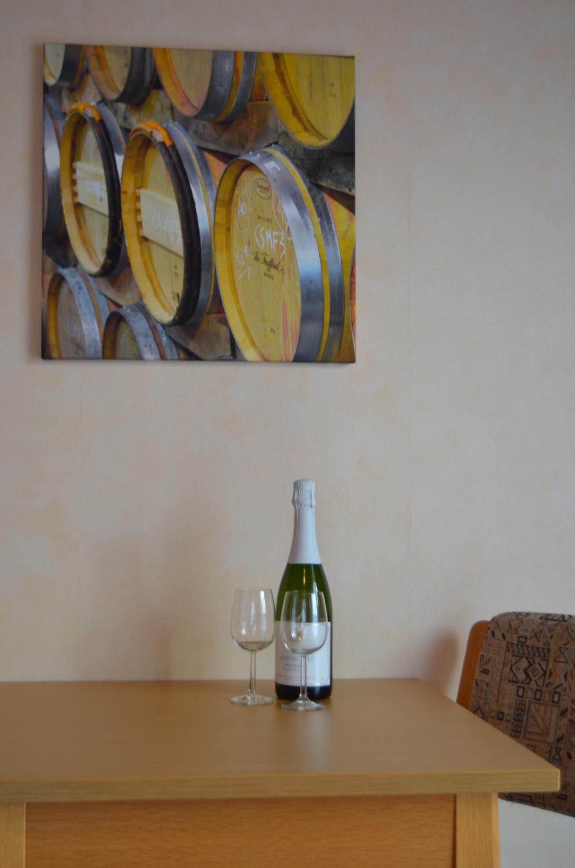 Une table avec une bouteille de vin et deux verres, un tableau représentant des tonneaux de vin accroché au mur et une chaise à motifs.