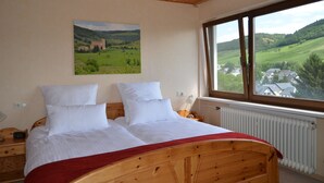 Ein Schlafzimmer mit einem hölzernen Bett, weißer Bettwäsche, einer roten Decke, einem Nachttisch, einer Lampe und einem Fenster mit Blick auf eine grüne Landschaft und ein Gebäude.