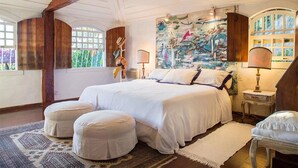 A spacious bedroom with a large bed, two ottomans, a painting, and a window with a view of greenery. - Village dos Ossos (Búzios)