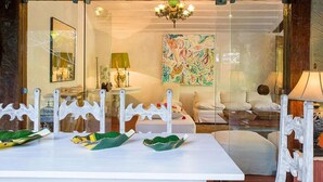 A dining area with a white table, chairs, and a glass partition. There is a chandelier above the table and a painting on the wall. - Village dos Ossos (Búzios)