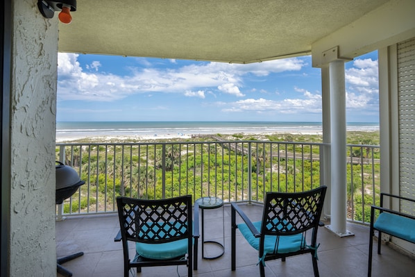 A balcony with two chairs and a table, overlooking a beach with a clear view of the ocean and distant shoreline.