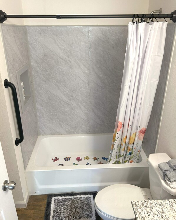 A shower area with a white shower curtain, a bathtub with a floral decal, a toilet, and a small rug.