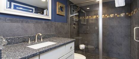A modern bathroom with a granite countertop, a white sink, and a glass-enclosed shower.